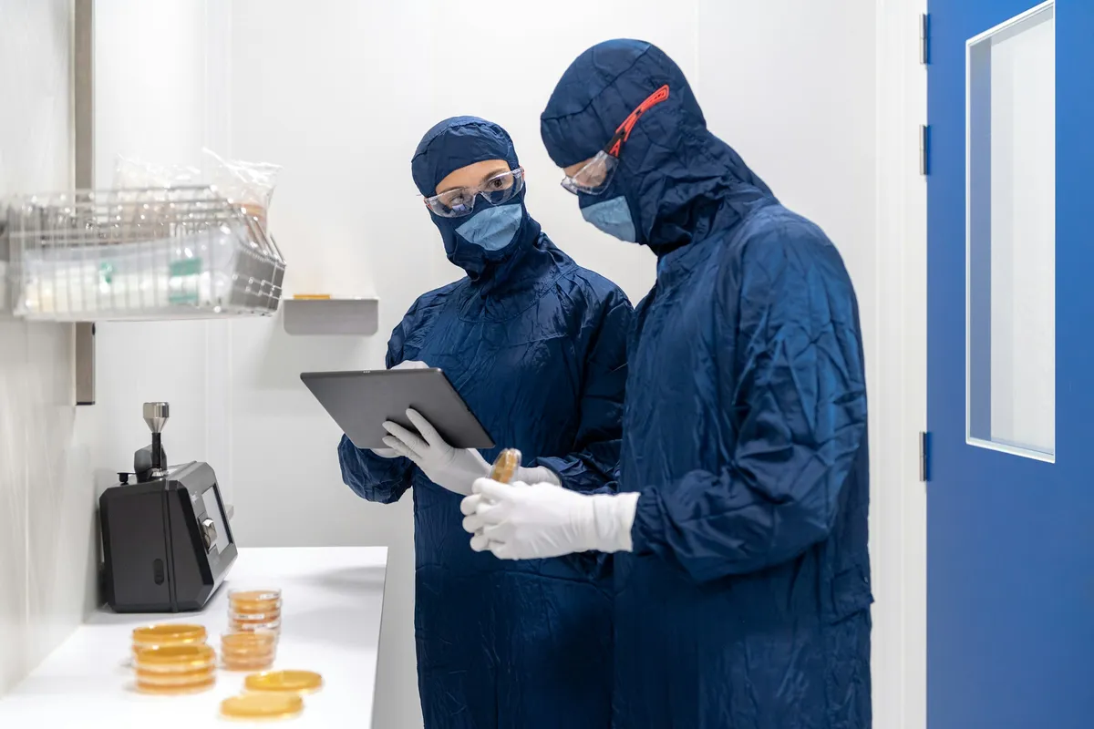 Dos operarios en trajes de cleanroom revisando placas de laboratorio con tablet.